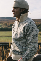 A man in profile wearing a cream and gray patterned knit quarter-zip sweater, a beige beanie, and a white collar, set against an outdoor rural landscape.