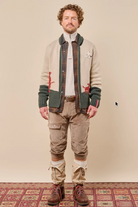 A man stands against a plain beige studio background wearing a cream knit cardigan with dark green ribbed trim and red embroidered details, layered over a brown plaid shirt, white turtleneck, tan cargo pants, and brown lace-up boots.