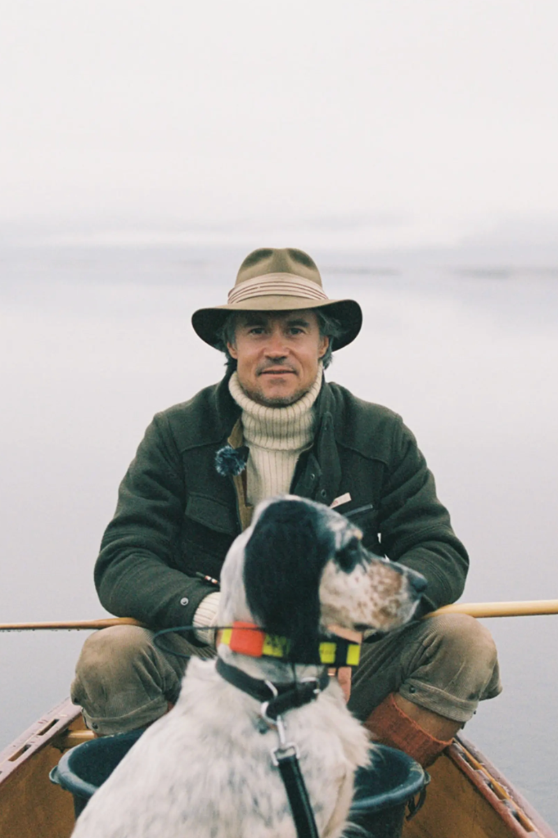 A man wearing a wide-brimmed hat, dark jacket, and cream turtleneck sits in a wooden boat with a black-and-white dog on a lake under an overcast sky.