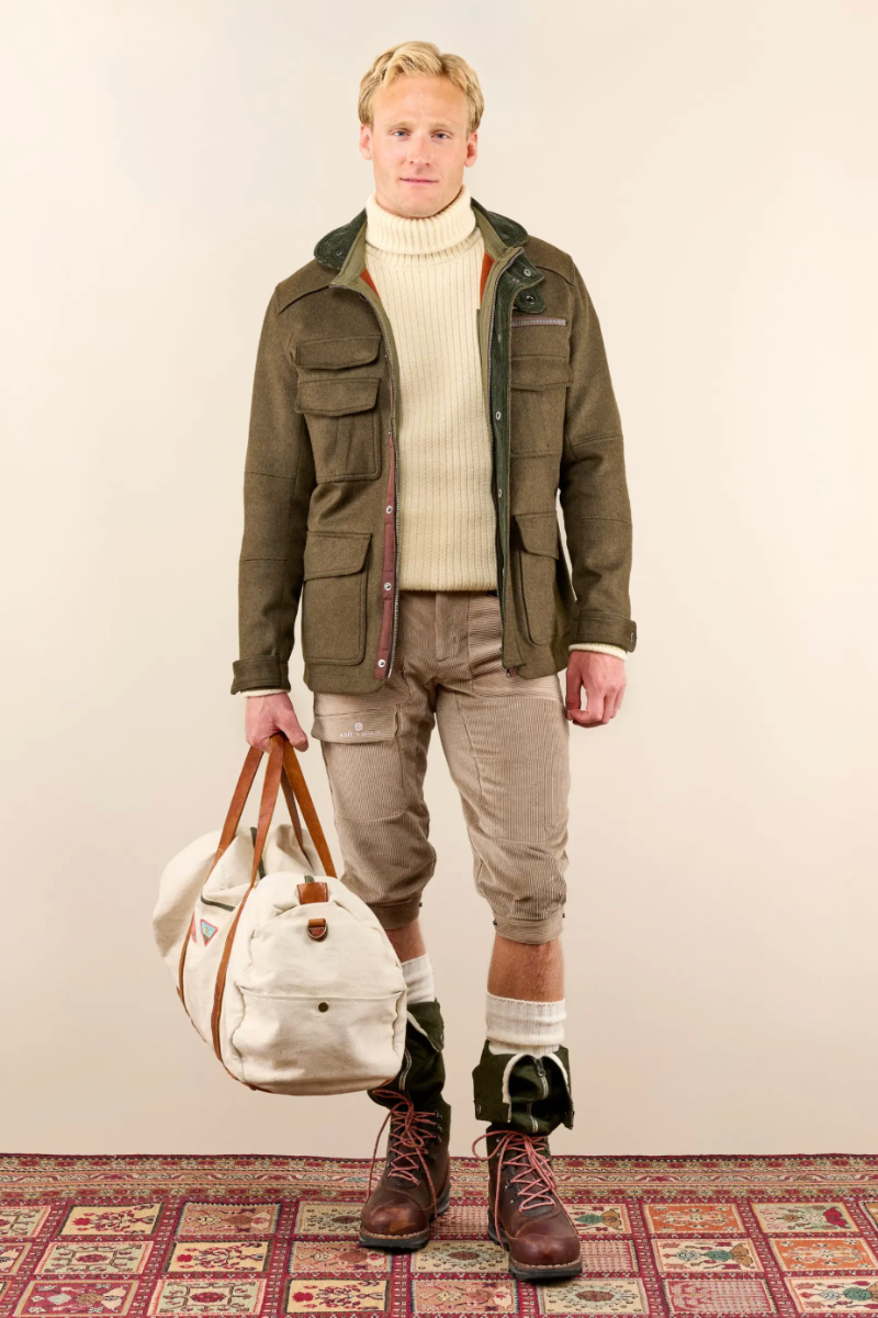 A man stands against a plain beige studio background wearing an olive green utility jacket over a cream turtleneck, beige cropped pants, white socks, and brown lace-up boots. He holds a large cream canvas duffel bag with brown handles.