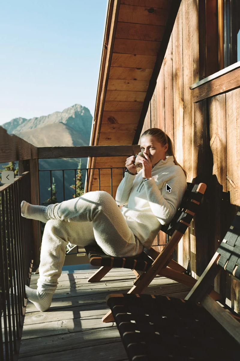 A woman relaxes in a wooden folding chair on a mountain chalet deck, wearing a matching cream fleece lounge set and holding a glass mug. The background features a rustic wooden wall and a scenic mountain landscape under a clear blue sky.