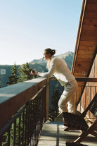 A woman in a cream-colored knit lounge set stands on a wooden deck of a cabin, looking out over a mountain and pine forest landscape.