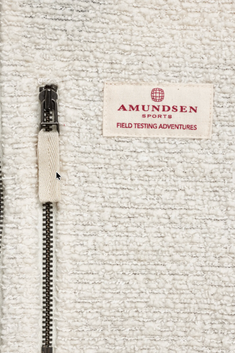 Close-up of a textured off-white fleece jacket featuring a black metal zipper with a matching beige fabric pull tab and a sewn-on brand label reading 'AMUNDSEN SPORTS FIELD TESTING ADVENTURES'.