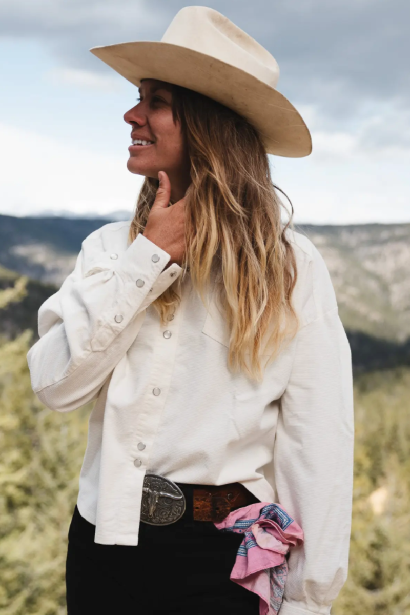 A woman wearing a cream cowboy hat, a cream western-style button-up shirt, and a black skirt with a large silver belt buckle, standing outdoors in a mountain landscape.