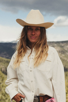 A woman wearing a cream-colored cowboy hat and matching long-sleeve western shirt, standing outdoors in front of a blurred mountain landscape.