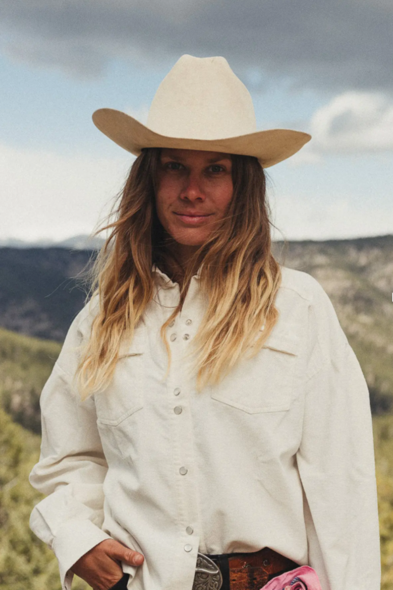 A woman wearing a cream-colored cowboy hat and matching long-sleeve western shirt, standing outdoors in front of a blurred mountain landscape.