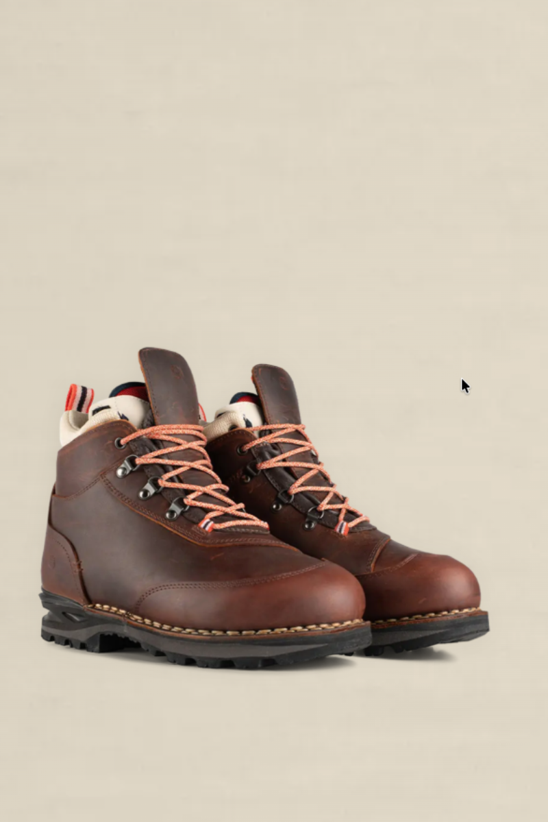 A pair of brown leather hiking boots with orange laces and a cream lining, shown against a solid beige background.