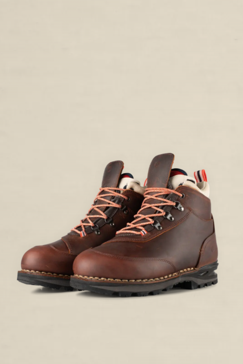 A pair of brown leather hiking boots with orange laces and a black sole, photographed on a clean, solid beige background.