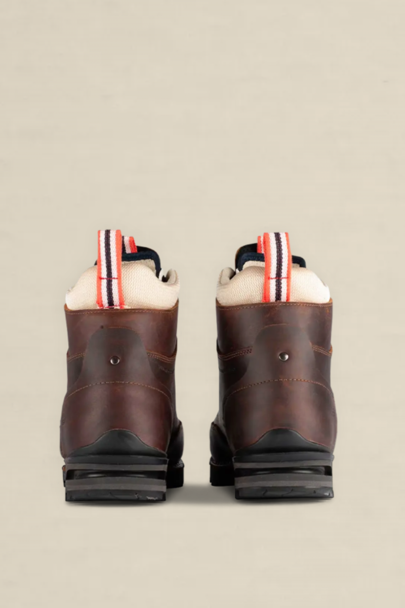 A pair of dark brown leather boots shown from the back, featuring a cream-colored fleece lining and a striped pull tab with red, white, and navy blue details, set against a solid light beige background.