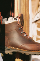 A close-up of a brown leather lace-up boot hanging vertically, with metal eyelets, orange-brown laces, and a rugged sole, set against a warm wooden and neutral background.