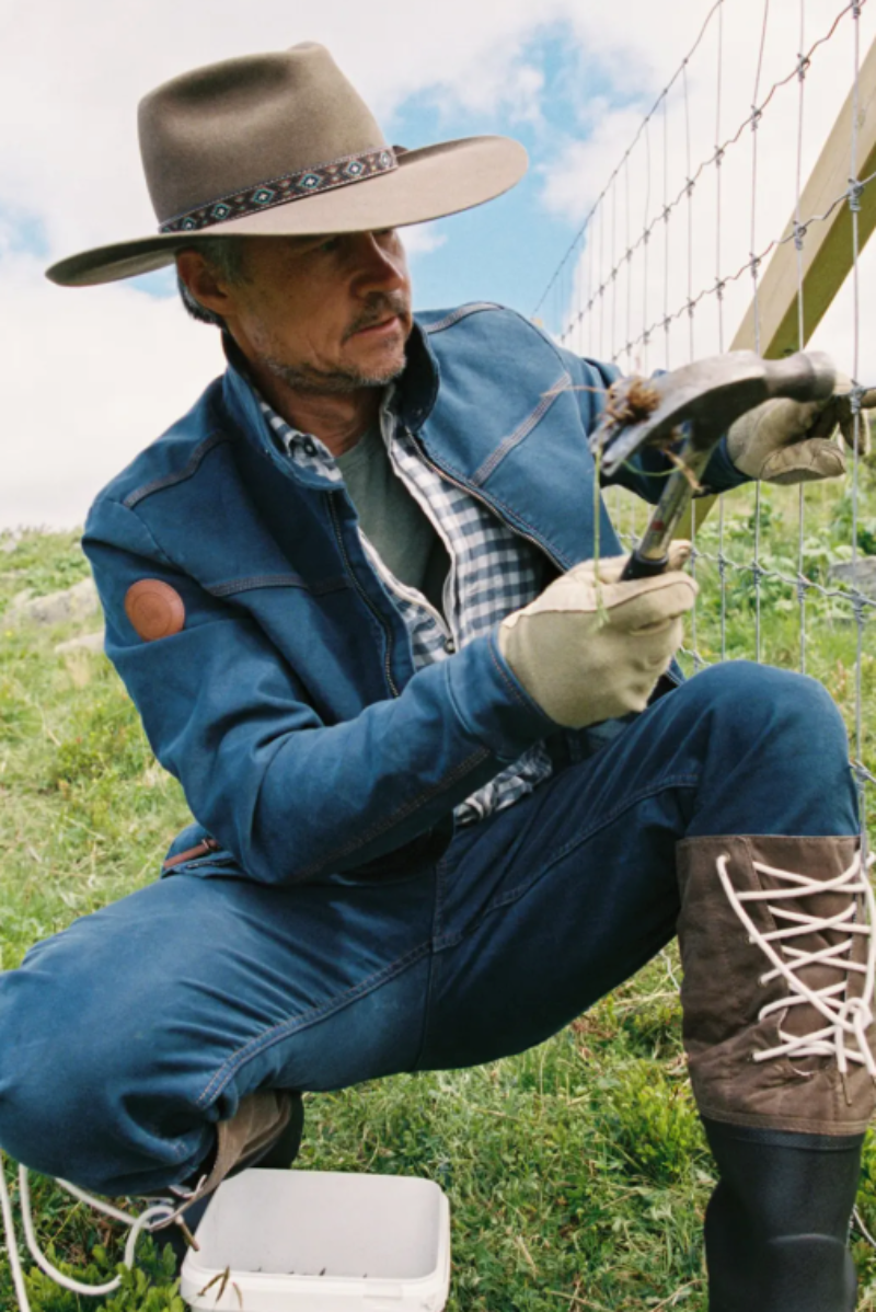 A man in western-style clothing, including a wide-brimmed hat, denim jacket, jeans, gloves, and lace-up boots, is crouching outdoors while working on a wire fence. He is holding a hammer and appears to be installing or repairing the fence in a grassy field under a blue sky.