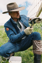 A man in western-style clothing, including a wide-brimmed hat, denim jacket, jeans, gloves, and lace-up boots, is crouching outdoors while working on a wire fence. He is holding a hammer and appears to be installing or repairing the fence in a grassy field under a blue sky.