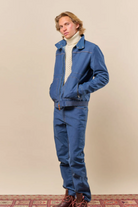 A man modeling a matching blue denim work jacket and pants set, styled with a cream cable-knit turtleneck sweater and brown boots, standing against a solid beige studio background.