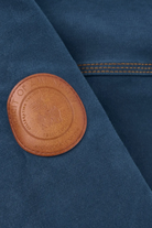 Close-up of a dark blue fabric garment featuring a circular brown leather patch on the chest with the text 'HONOR OF AMONGST ANTIQUED GILTY'.