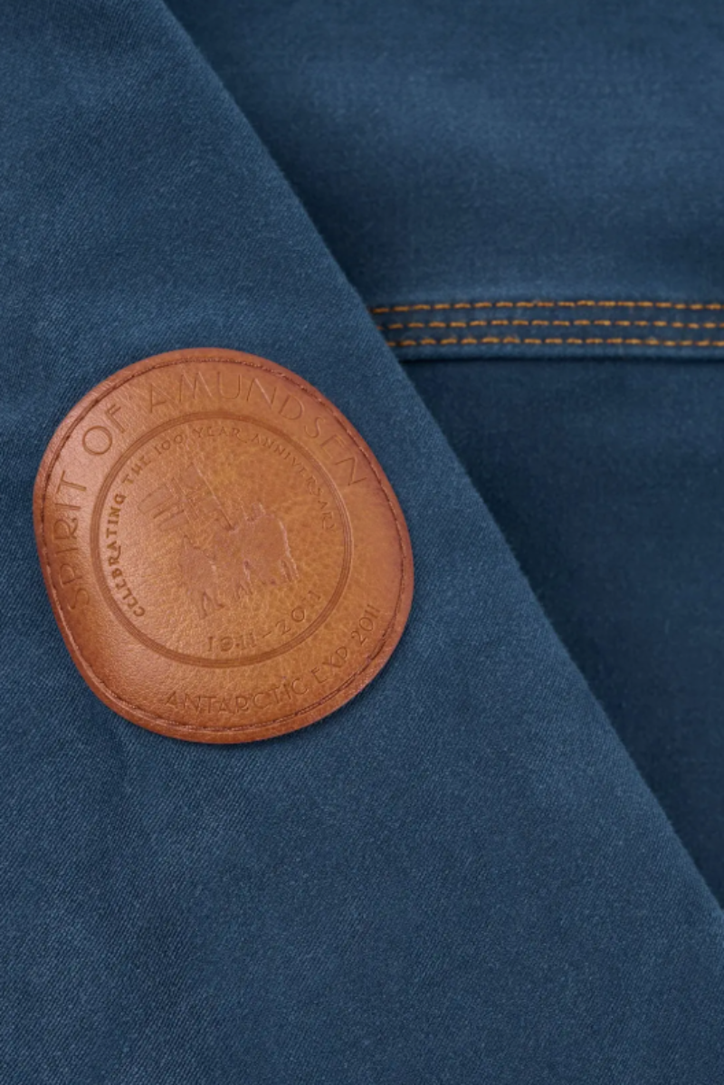 Close-up of a dark blue fabric garment featuring a circular brown leather patch on the chest with the text 'HONOR OF AMONGST ANTIQUED GILTY'.