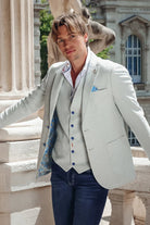 Man in a light gray suit with blue buttons standing outdoors near classical architecture.