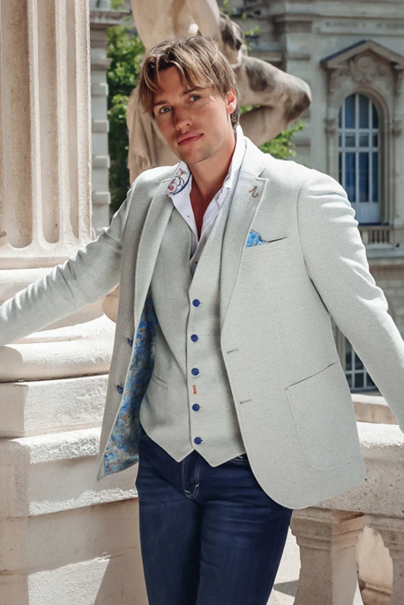 Man in a light gray suit with blue buttons standing outdoors near classical architecture.