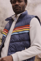 Man wearing a blue puffer vest with colorful stripes in a natural setting
