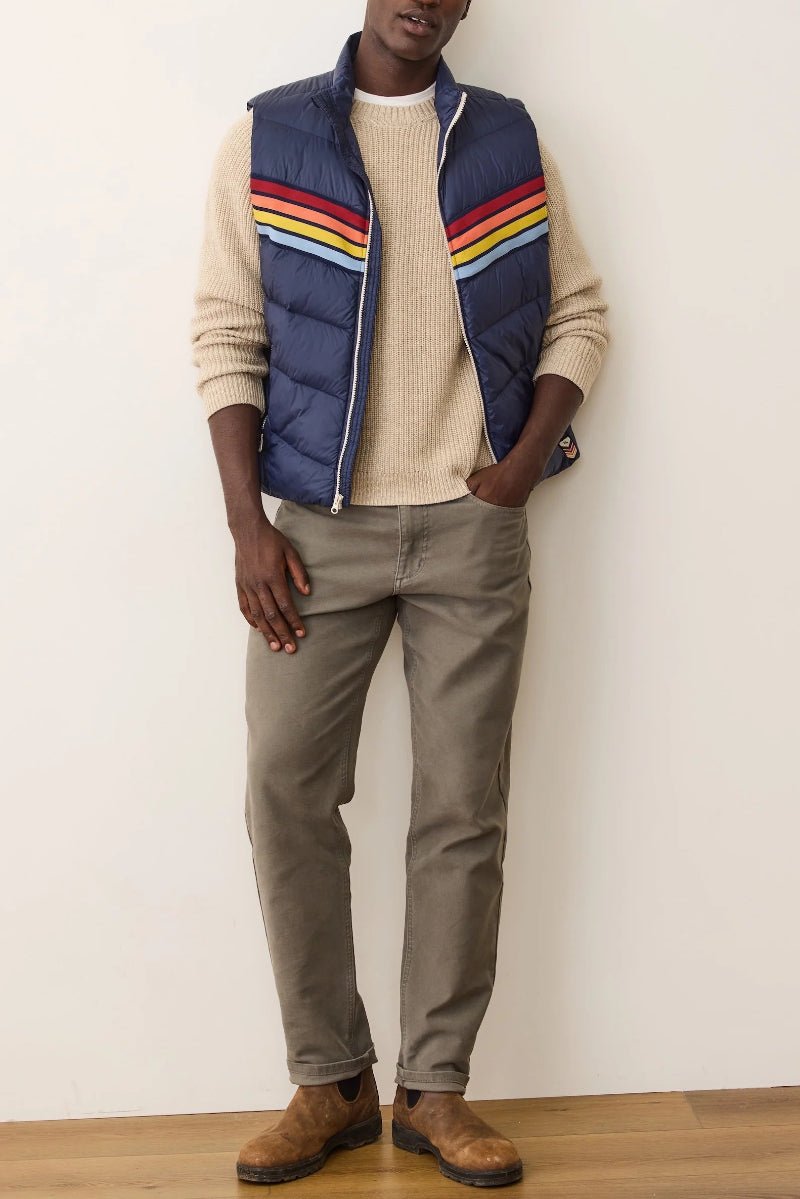 Person wearing a blue puffer vest with colorful stripes, beige sweater, and gray pants against a white wall.