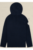A dark navy blue textured hooded jacket shown from the back, featuring a circular patch on the left sleeve, set against a solid light beige background.
