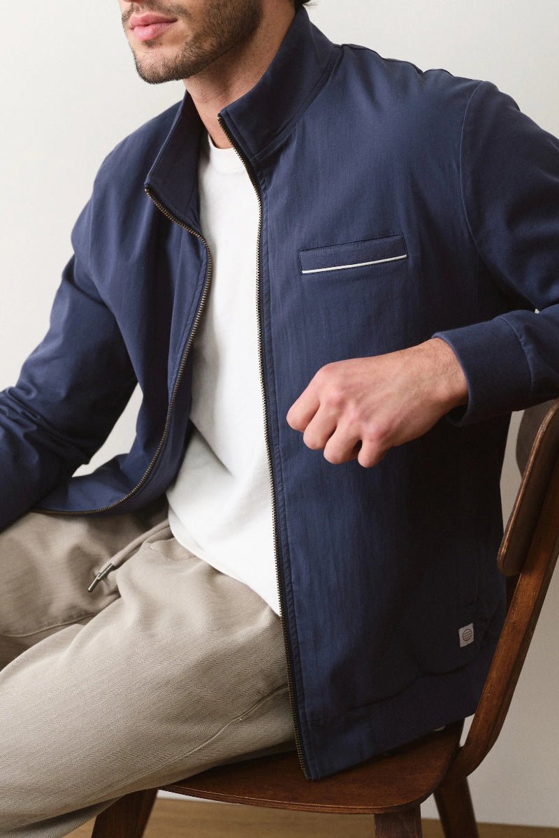Man wearing a navy jacket sitting on a wooden chair against a plain background
