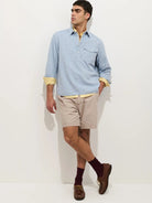 Man wearing a light blue shirt with yellow cuffs and trim, beige shorts, and brown shoes on a white background.