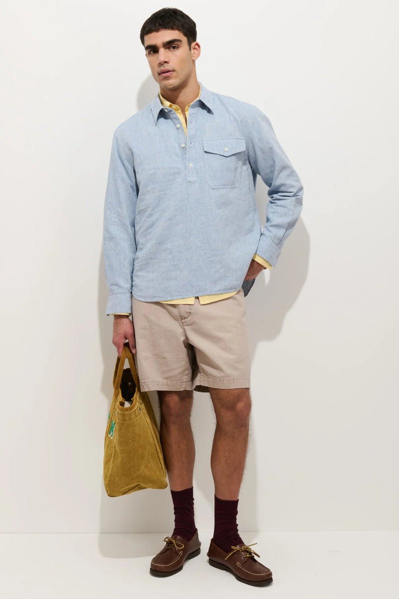 Man wearing a light blue long-sleeve shirt, beige shorts, and brown shoes holding a yellow bag against a white background.