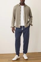 Man wearing a beige shirt, white t-shirt, blue pants, and white sneakers against a plain background