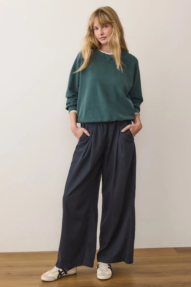 Woman wearing a green sweater and dark pants against a white wall.