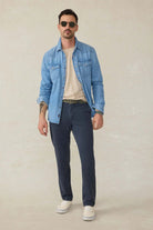 Man wearing a light blue denim jacket, white shirt, dark blue pants, and sunglasses on a beige background