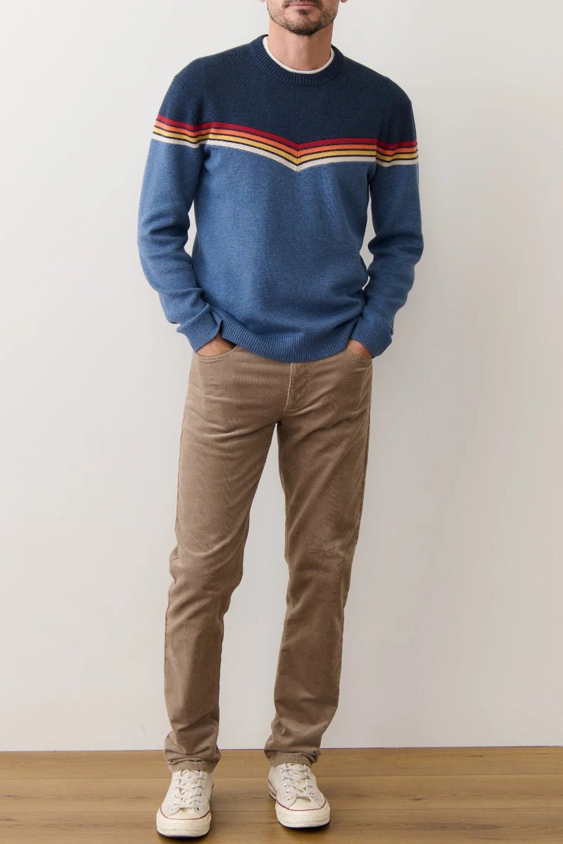 Man wearing a blue sweater with colorful stripes and brown pants against a plain background