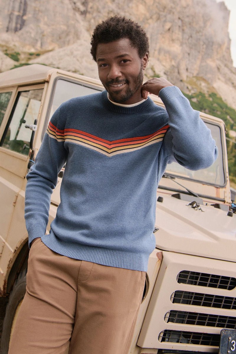 Man in a blue sweater with colorful stripes standing next to a vintage SUV in a mountainous area.