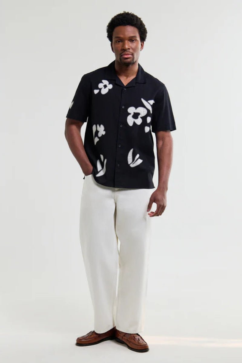 Man wearing a black shirt with white floral patterns and white pants on a white background