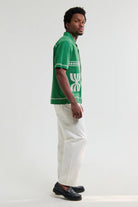 Man wearing a green polo shirt with a logo and white pants on a light gray background