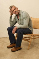 Man sitting on a wooden chair wearing a light green sweater and dark jeans.