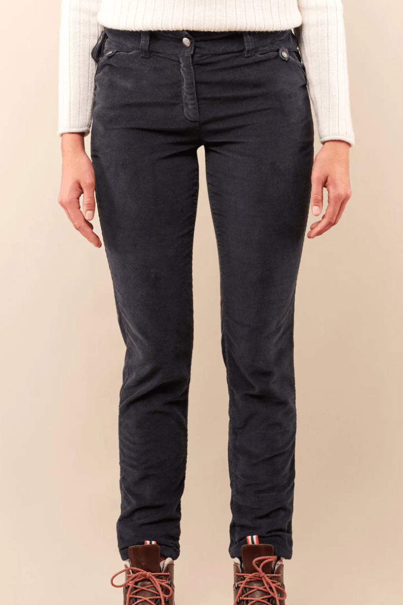 A person wearing slim-fit dark navy jeans, a white ribbed long-sleeve top, and brown lace-up boots, standing against a plain beige studio background.
