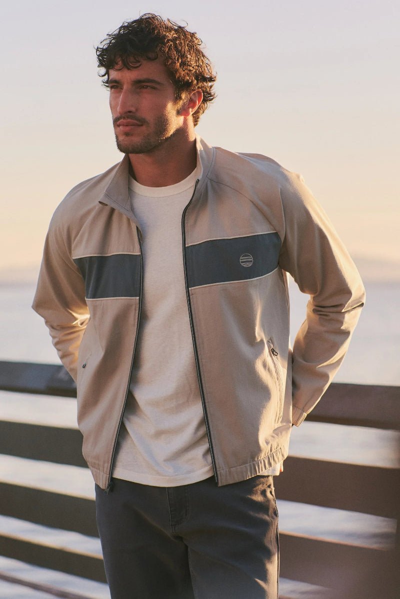 Man wearing a beige jacket with a blue patch, standing by a body of water.