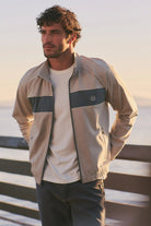 Man wearing a beige jacket with a blue patch, standing by a body of water.