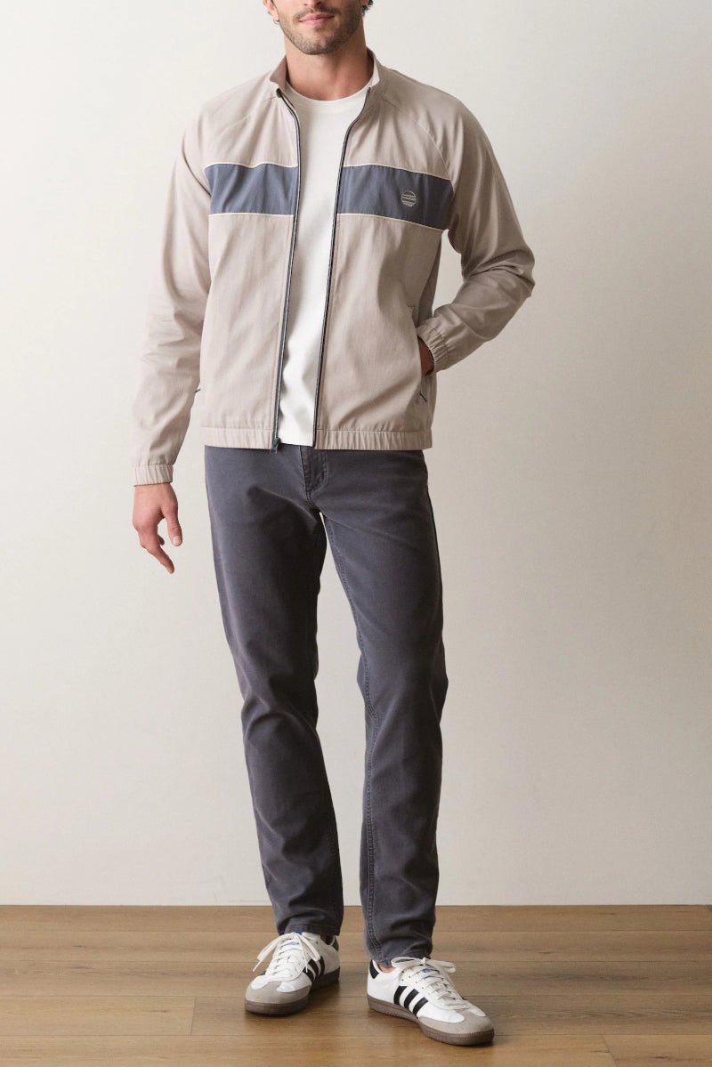 Man wearing a beige jacket with a logo, white shirt, gray pants, and Adidas shoes on a plain background