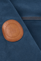 Close-up of a dark blue fabric garment featuring a circular brown leather patch on the chest with the text 'HONOR OF AMONGST ANTIQUED GILTY'.