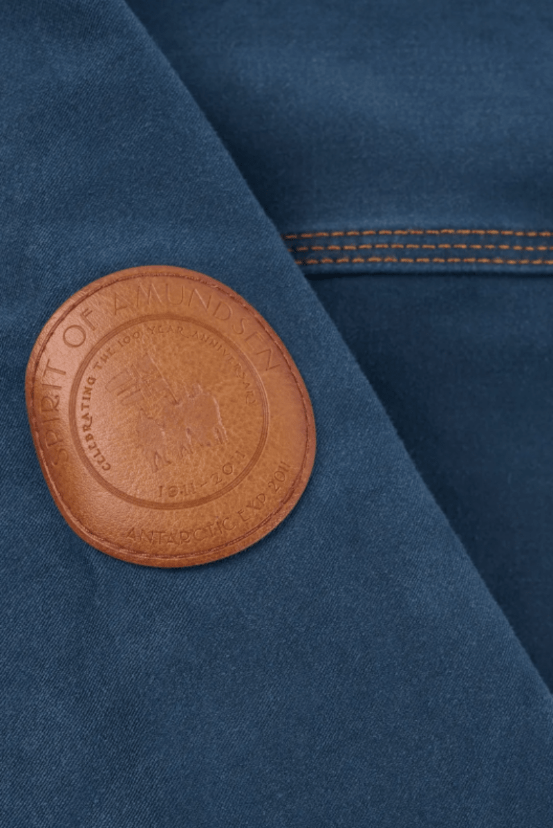 Close-up of a dark blue fabric garment featuring a circular brown leather patch on the chest with the text 'HONOR OF AMONGST ANTIQUED GILTY'.
