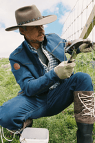 A man in western-style clothing, including a wide-brimmed hat, denim jacket, jeans, gloves, and lace-up boots, is crouching outdoors while working on a wire fence. He is holding a hammer and appears to be installing or repairing the fence in a grassy field under a blue sky.