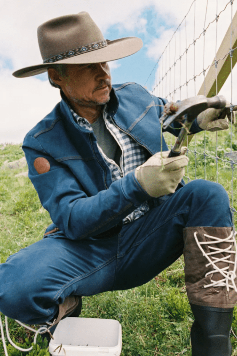 A man in western-style clothing, including a wide-brimmed hat, denim jacket, jeans, gloves, and lace-up boots, is crouching outdoors while working on a wire fence. He is holding a hammer and appears to be installing or repairing the fence in a grassy field under a blue sky.