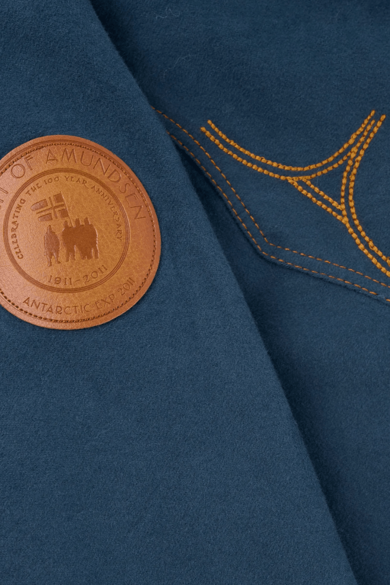 Close-up of a dark teal wool-blend jacket featuring a circular brown leather patch on the chest with embossed text and a gold embroidered geometric pattern.