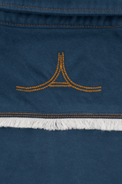 Close-up of a dark blue suede or nubuck textile with orange contrast stitching, a small orange embroidered logo motif, and a white fringe trim.
