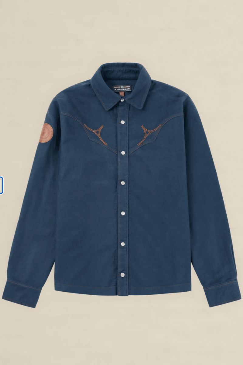 A dark blue denim western-style shirt jacket with brown embroidered antlers across the chest, a brown circular patch on the right sleeve, and white buttons, shown against a light beige background.