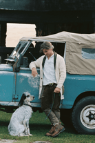 A man in a cream cardigan and brown boots leans against a vintage blue off-road vehicle next to a white and black spotted dog on a grassy outdoor setting.