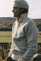A man in profile wearing a cream and gray patterned knit quarter-zip sweater, a beige beanie, and a white collar, set against an outdoor rural landscape.