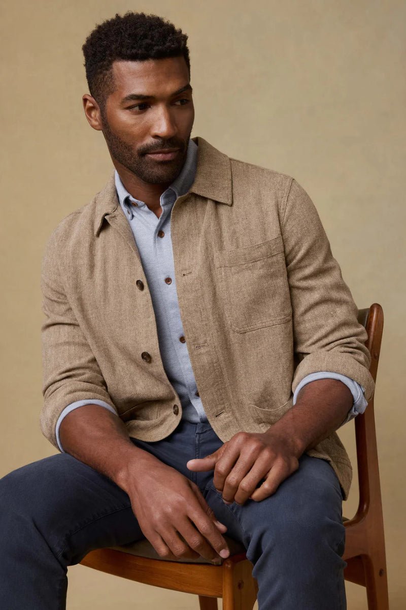 Man sitting on a wooden chair wearing a beige jacket over a light blue shirt.