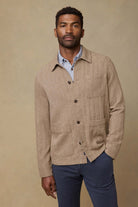 Man wearing a beige jacket over a blue shirt and dark pants on a beige background